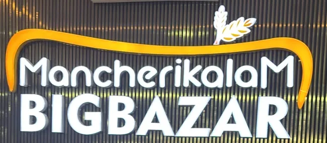 The lit MancherikalaM BigBazar sign — white letters under a golden arc, on a dark slatted wall